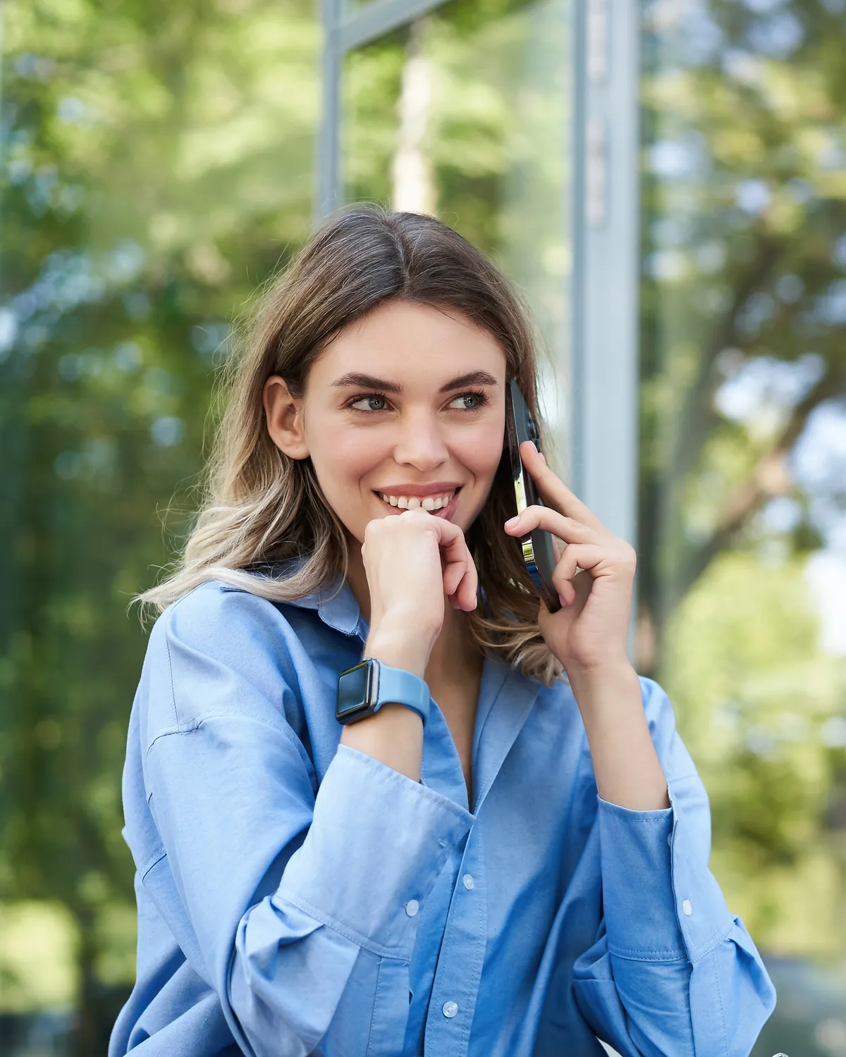 A woman on a phone call at her laptop, glass and trees behind